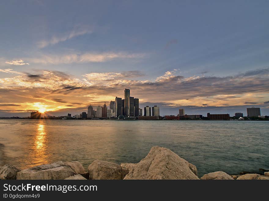 Skyscrapers and buildings viewed from the rocky banks of a river (opposite) at sunset with the sun creating an orange glow and above it pale blue sky. Skyscrapers and buildings viewed from the rocky banks of a river (opposite) at sunset with the sun creating an orange glow and above it pale blue sky.