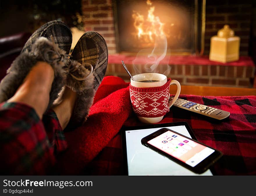 A person with slippers relaxing by the fireplace with a cup of coffee and smartphone. A person with slippers relaxing by the fireplace with a cup of coffee and smartphone.