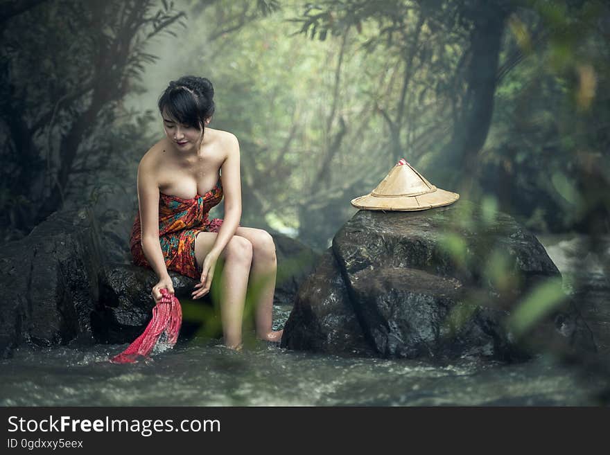 Beautiful young Asian woman sat on rocks in forest washing clothes in stream. Beautiful young Asian woman sat on rocks in forest washing clothes in stream.