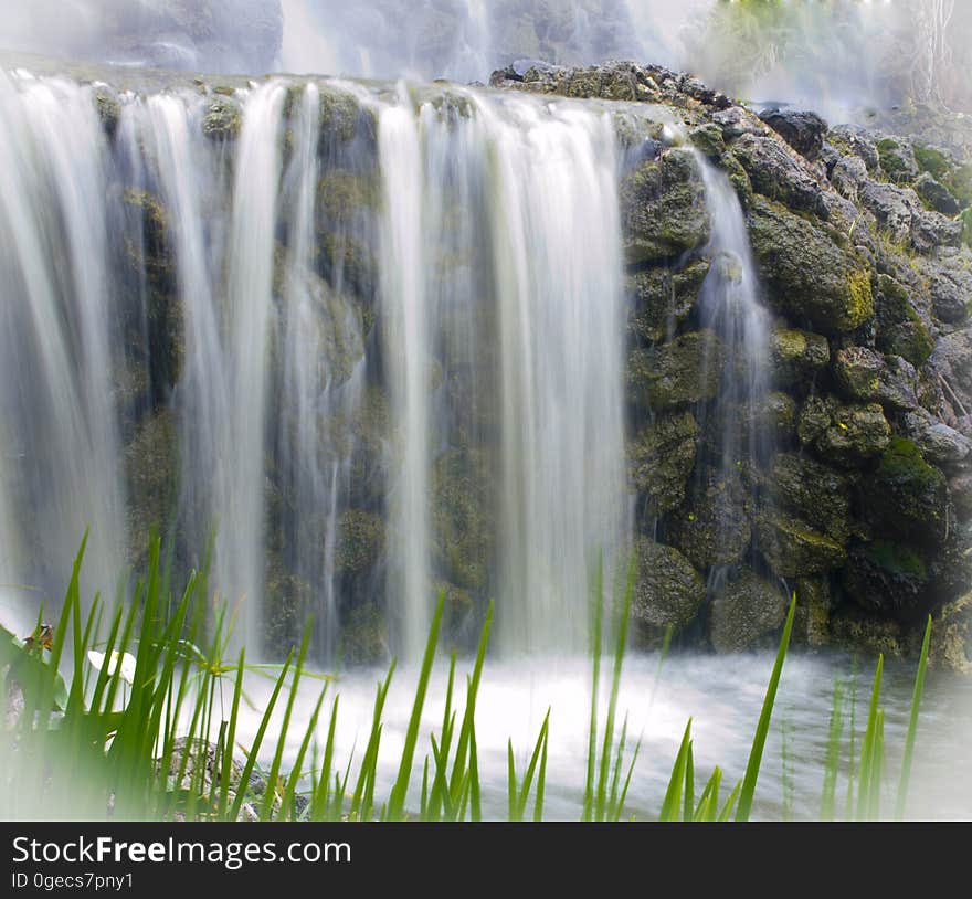 Time lapse scenic view of waterfall over rocky cliff with blades of grass in foreground.