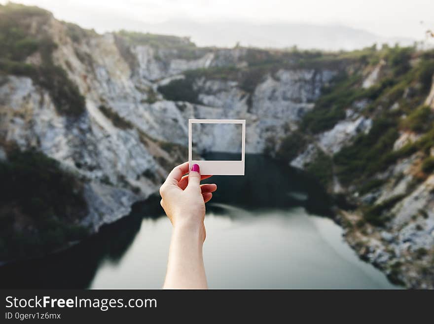 A person holding a polaroid photo that captures the cliffs in front of it. A person holding a polaroid photo that captures the cliffs in front of it.