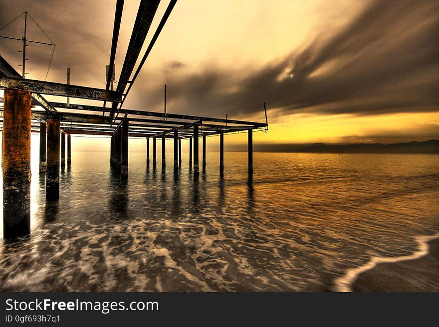 Pier structure without the usual wooden boards lit by an orange and yellow light of the sunset with the tide coming in and the sea decorated by a foam pattern. Pier structure without the usual wooden boards lit by an orange and yellow light of the sunset with the tide coming in and the sea decorated by a foam pattern.