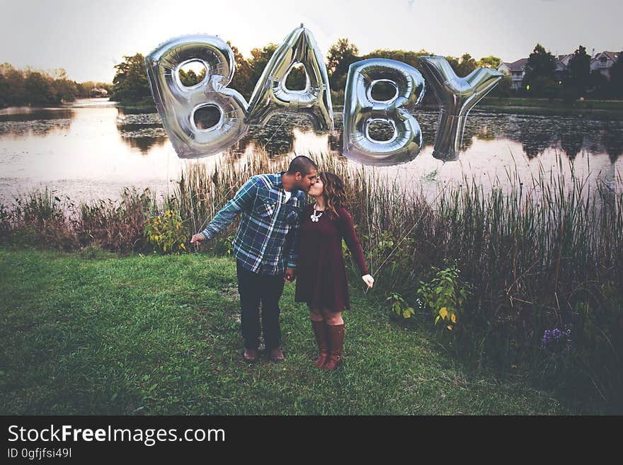 Loving couple standing beside a river kissing passionately with rubber balloons over their heads spelling "baby" in uppercase letters ( 3D shapes) possibly indicating the desire for a child. Loving couple standing beside a river kissing passionately with rubber balloons over their heads spelling "baby" in uppercase letters ( 3D shapes) possibly indicating the desire for a child.