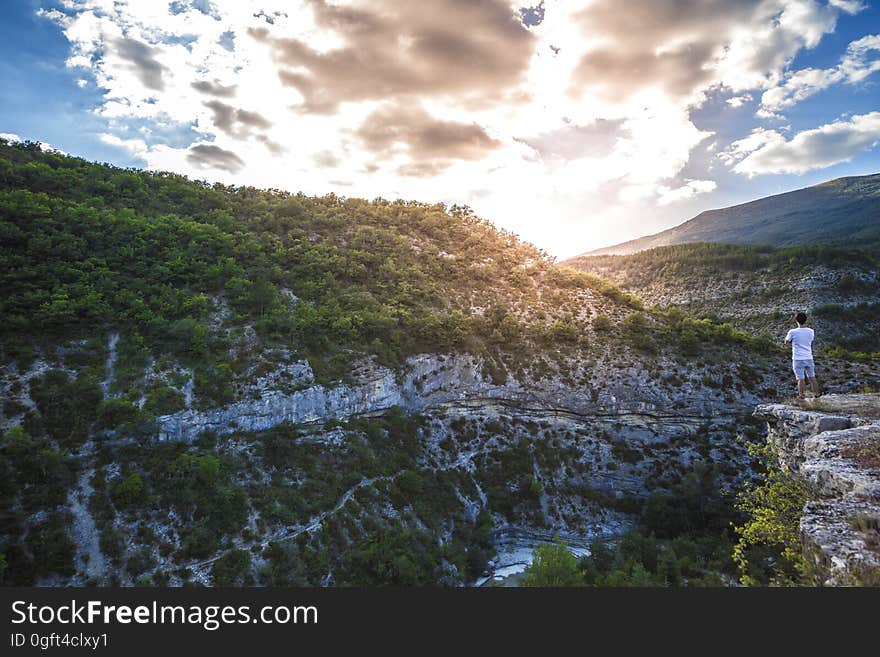 Man in White Shirt Standing on Top of Mountain Cliff Under Cumulus Clouds