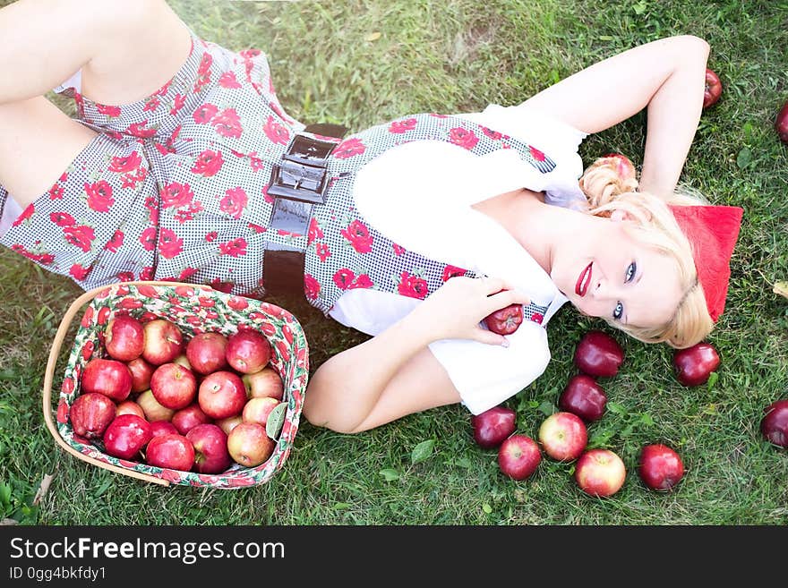 Woman in Gray and White Rose Print Onesies Laying on Grass Beside Red Apple Fruits