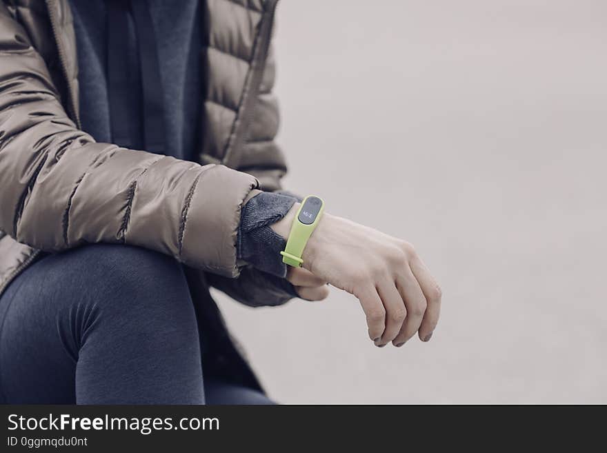 Closeup of man (face not shown) in quilted jacket wearing pale green colored electronic watch on his wrist and blue jeans with the watch maybe capable of monitoring physiological features (pulse, distance walked), fawn background. Closeup of man (face not shown) in quilted jacket wearing pale green colored electronic watch on his wrist and blue jeans with the watch maybe capable of monitoring physiological features (pulse, distance walked), fawn background.