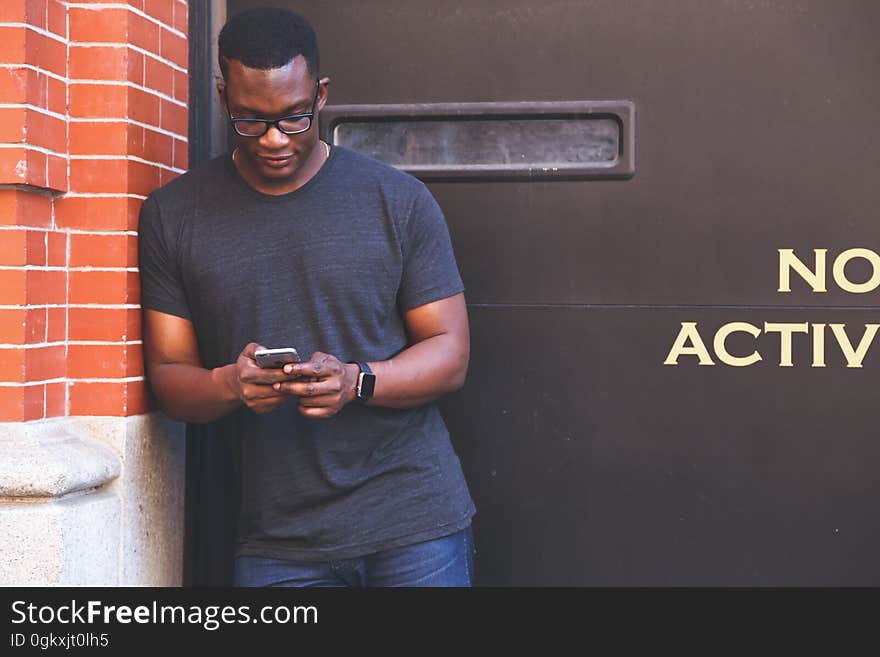 Man in Grey Crew Neck T Shirt Leaning on Brown Brick Wall Holding Smartphone