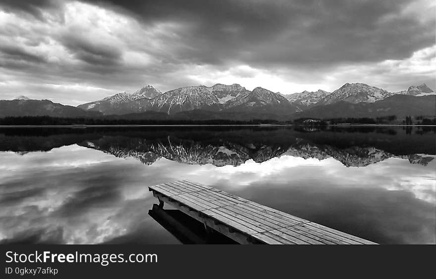 Black and white panoramic view of mountains reflecting on lake with pier in foreground.