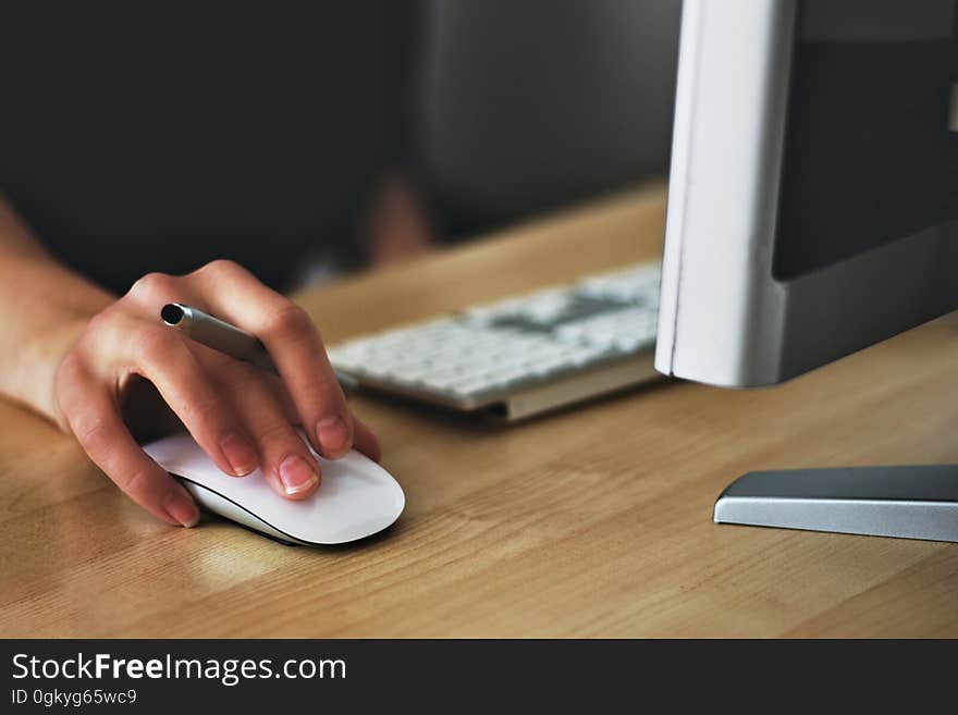 Selective focus on woman's hand and fingers on mouse next to computer keyboard and display, also holding pen between forefinger and second finger. Selective focus on woman's hand and fingers on mouse next to computer keyboard and display, also holding pen between forefinger and second finger.