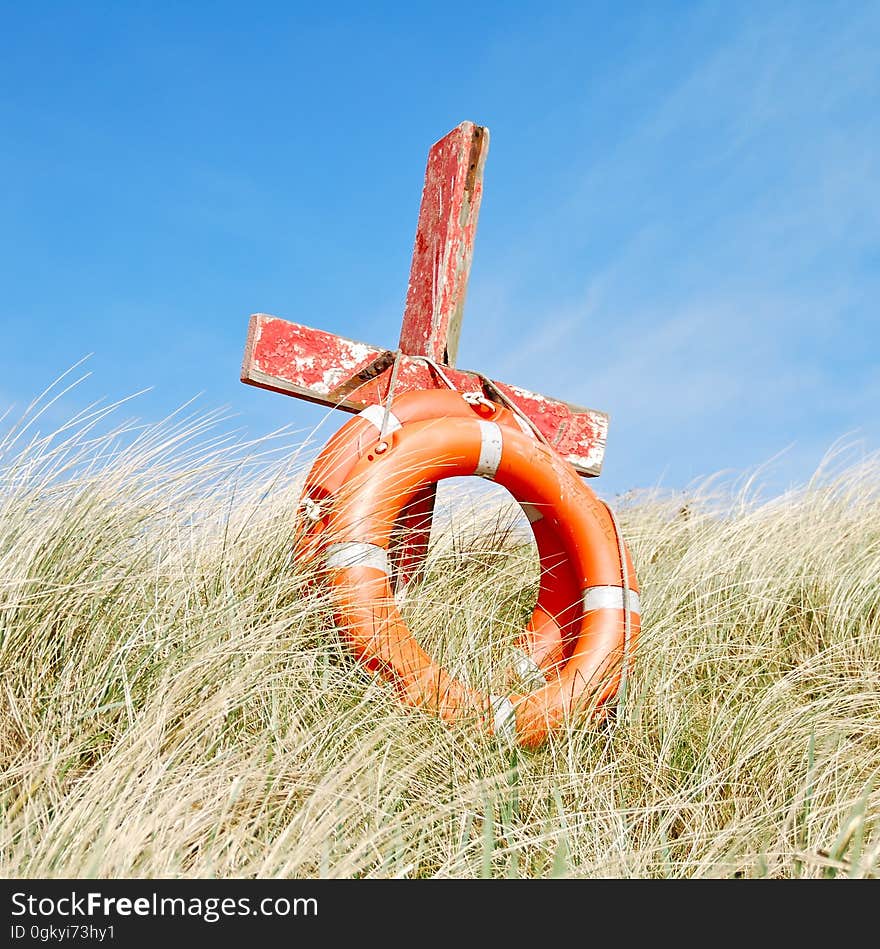 Two red life buoys tied to a wooden cross in coarse bleached grasses planted close to sand dunes and the seashore, perfect blue sky.