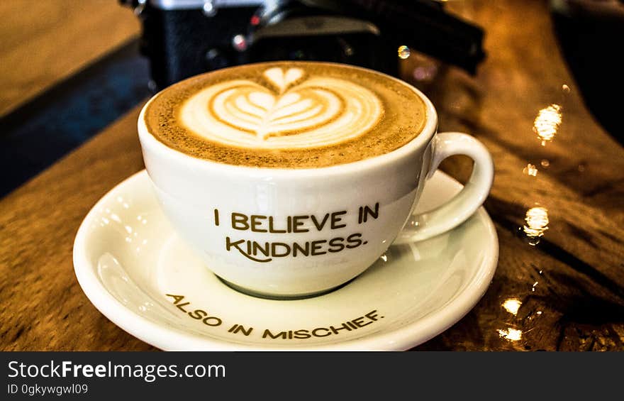 White chine cup and saucer with message I Believe in Kindness and Also in Mischief with latte coffee drink. White chine cup and saucer with message I Believe in Kindness and Also in Mischief with latte coffee drink.