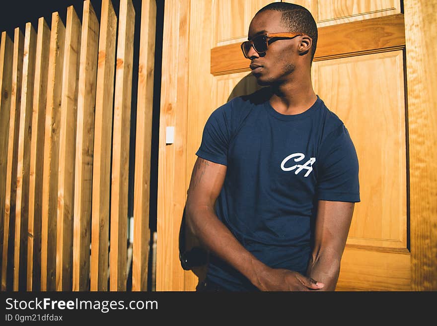 Portrait of African man in blue t-shirt wearing sunglasses leaning against wooden wall in sunshine. Portrait of African man in blue t-shirt wearing sunglasses leaning against wooden wall in sunshine.