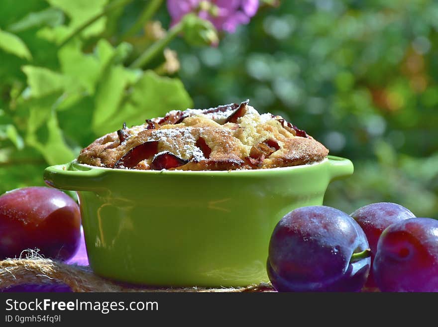 Baked cake with fresh plumbs on table outdoors, green leaves and flowers in background.