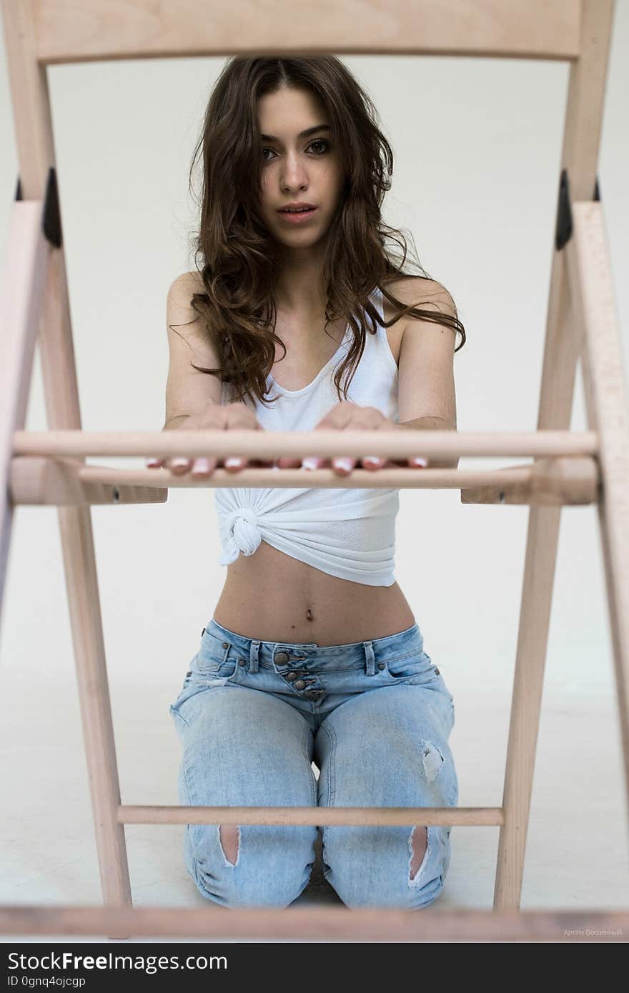 Brunette girl in white top and jeans sitting on his knees in front of the wooden chair