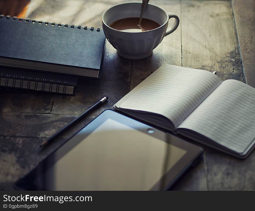 Still life with tablet or e-reader on rustic table with blank journal and cup of coffee. Still life with tablet or e-reader on rustic table with blank journal and cup of coffee.