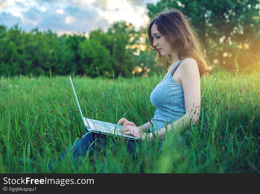 Woman sitting on a green meadow with trees on the background of sunset with clouds. Working or studying on laptop wireless. The concept of education with nature