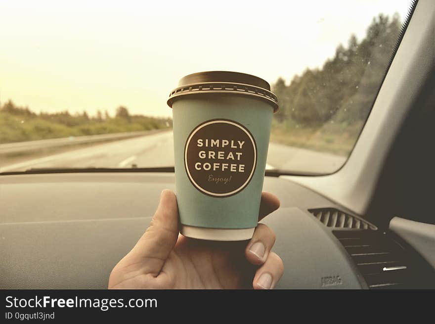 Hand holding disposable coffee cup inside car on road. Hand holding disposable coffee cup inside car on road.