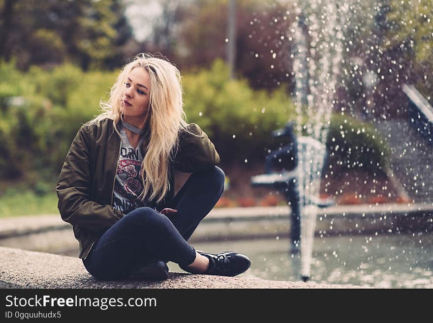 Model with long blond hair poses wearing jeans and leather jacket and sits cross legged beside a pool with a water fountain, blurred bushes and trees in the background. Model with long blond hair poses wearing jeans and leather jacket and sits cross legged beside a pool with a water fountain, blurred bushes and trees in the background.