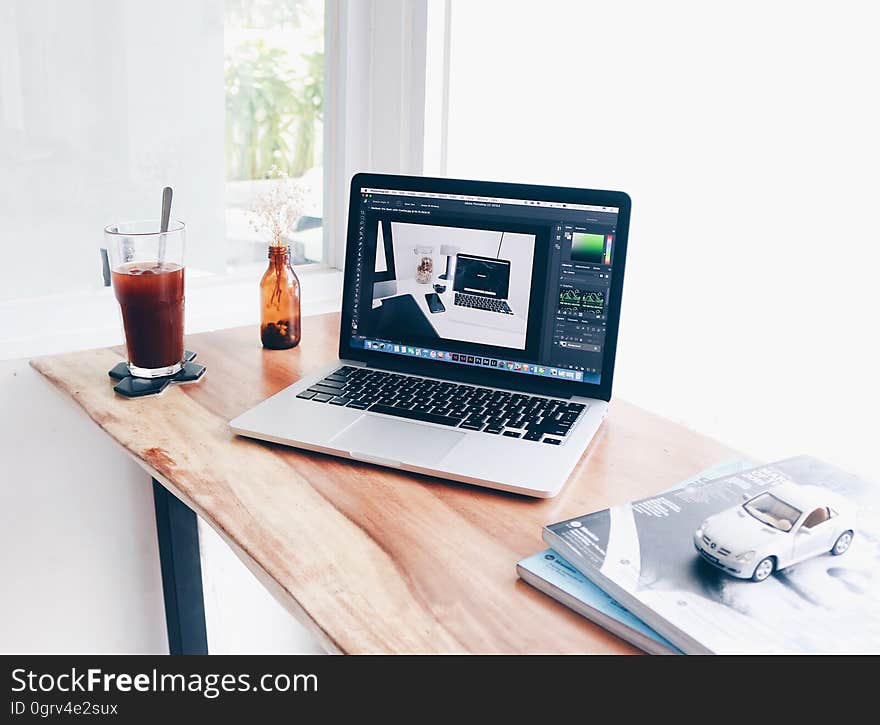 Wooden desk with laptop computer, drink, bottle, books and model car placed on it, white walls and blurred window. Wooden desk with laptop computer, drink, bottle, books and model car placed on it, white walls and blurred window.