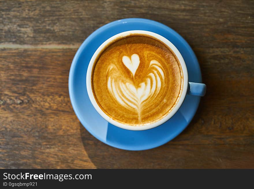 Overhead view of heart shape in foam of coffee in blue china cup with saucer on wood. Overhead view of heart shape in foam of coffee in blue china cup with saucer on wood.