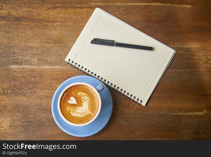 Overhead view of coffee on wooden table with decorative heart shaped next to notepad and pen. Overhead view of coffee on wooden table with decorative heart shaped next to notepad and pen.