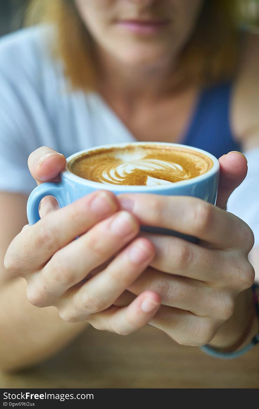 Closeup of two hands of a woman wrapped around a blue cup or mug of coffee (latte) with a partial blurred face behind. Closeup of two hands of a woman wrapped around a blue cup or mug of coffee (latte) with a partial blurred face behind.