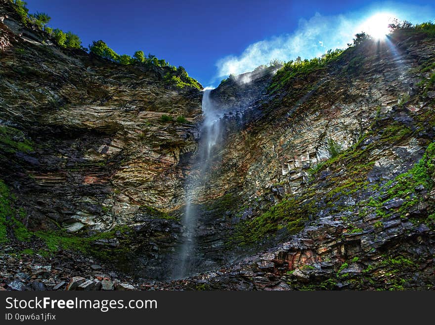 An impressive waterfall down a rocky cliff with a fringe of trees on top and green moss down below, blue sky and white cloud above.