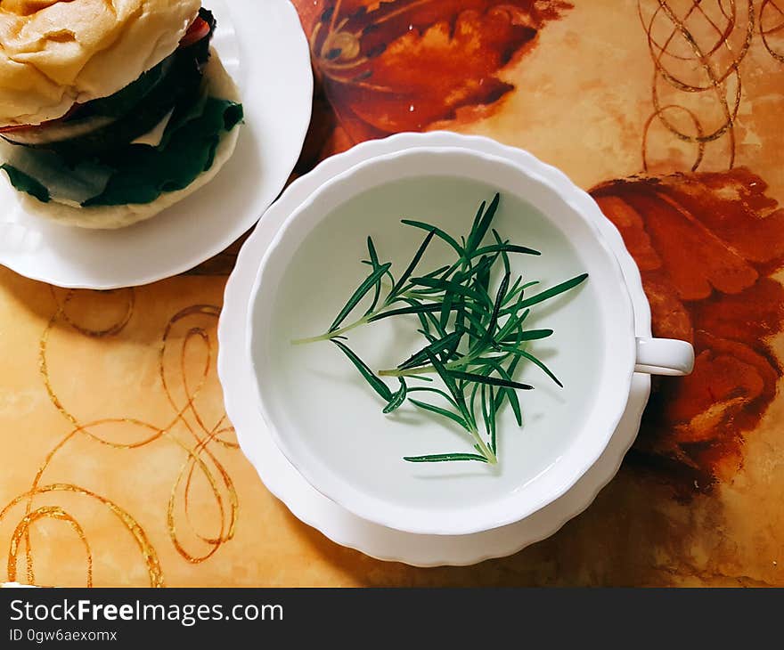 Cup and saucer with green herbs in clear broth with hamburger on white china plate. Cup and saucer with green herbs in clear broth with hamburger on white china plate.