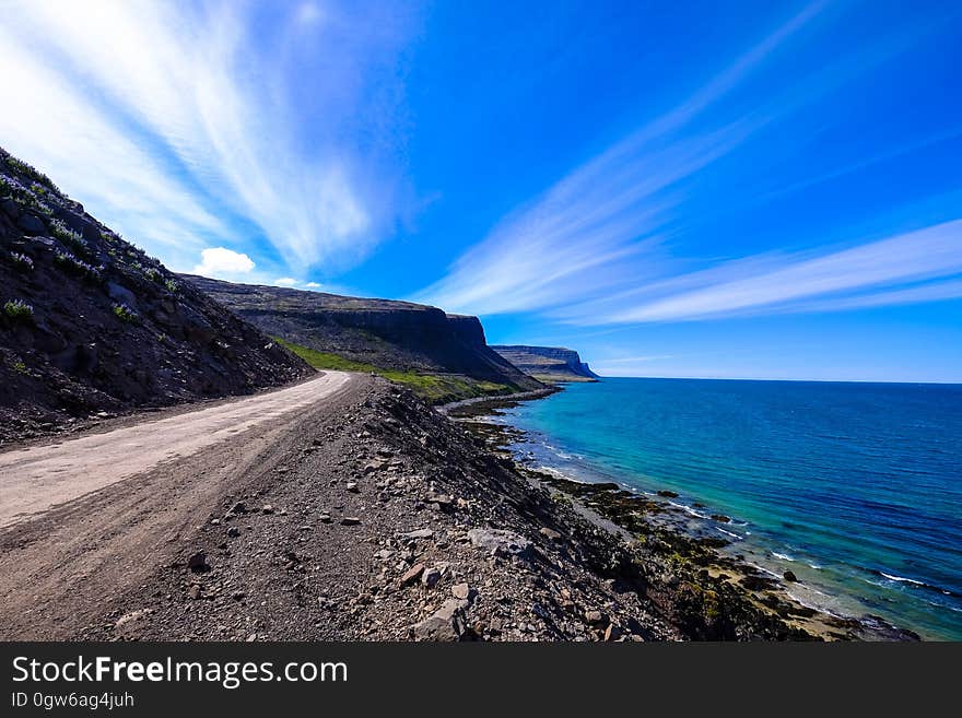 Rough coast road beside an aquamarine sea with dramatic sky with clouds in divergent white streaks. Rough coast road beside an aquamarine sea with dramatic sky with clouds in divergent white streaks.