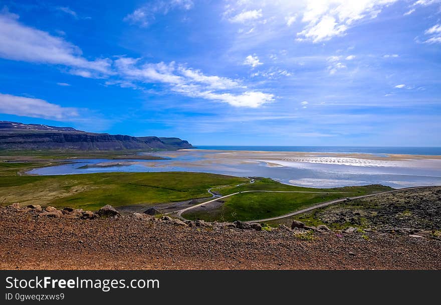 View from a rough road (or car park) over fields to a blue stretch of sea at low tide with sand banks showing and beyond bright blue sky and fluffy clouds. View from a rough road (or car park) over fields to a blue stretch of sea at low tide with sand banks showing and beyond bright blue sky and fluffy clouds.