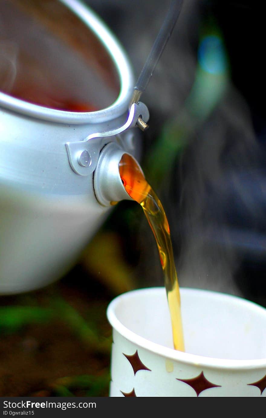 Pouring breakfast tea from a metal pot into a (plastic) patterned cup, blurred background.