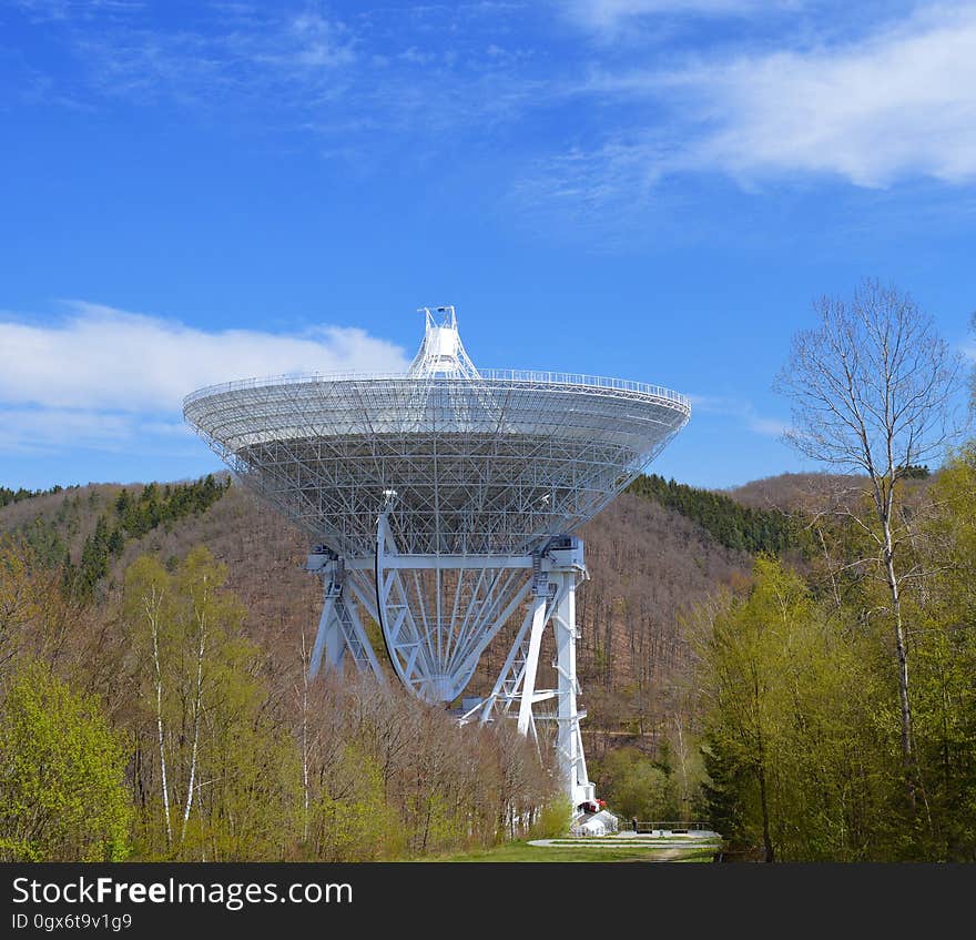 Observatory with large complicated metal dish and support structure surrounded by trees and forest, bright blue sky with a few clouds. Observatory with large complicated metal dish and support structure surrounded by trees and forest, bright blue sky with a few clouds.