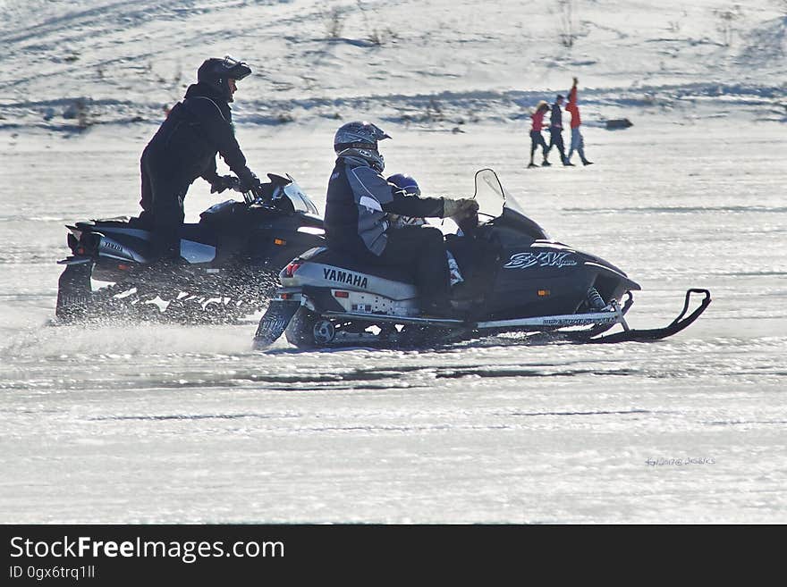 A pair of snow mobiles with people riding them in the snow. A pair of snow mobiles with people riding them in the snow.