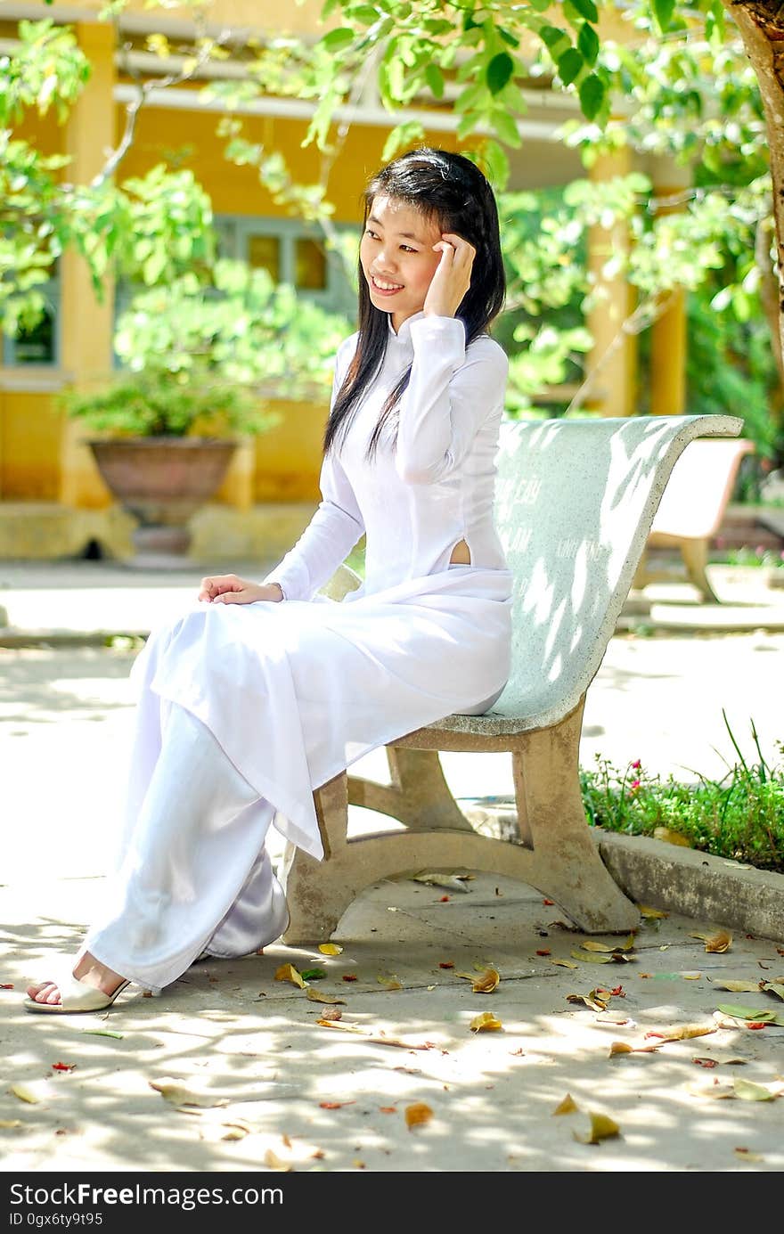 Young smiling Asian girl wearing white dress sitting in the park posing, blurred background. Young smiling Asian girl wearing white dress sitting in the park posing, blurred background.