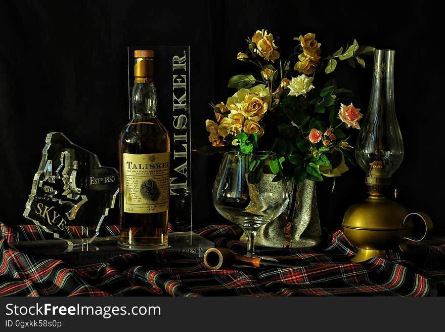 Table covered in tartan cloth with bottle of Talisker whisky vase of cream and pink roses, oil lamp, glass and map of Isle of Skye in glass, dark background. Table covered in tartan cloth with bottle of Talisker whisky vase of cream and pink roses, oil lamp, glass and map of Isle of Skye in glass, dark background.