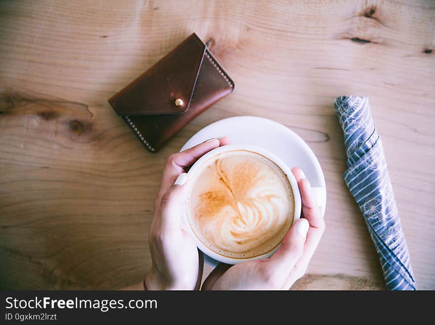 Two hands holding cup of coffee on a knotty wooden table with purse (or wallet) nearby and with cutlery wrapped in napkin. Two hands holding cup of coffee on a knotty wooden table with purse (or wallet) nearby and with cutlery wrapped in napkin.