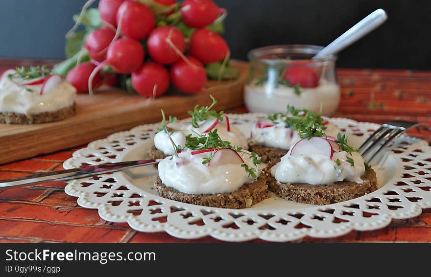 Four slices of baked bread with white sauce and garnishment on a white plate placed on a table, with a single bread and sauce, radishes, and a bowl of sauce in the background. Four slices of baked bread with white sauce and garnishment on a white plate placed on a table, with a single bread and sauce, radishes, and a bowl of sauce in the background.