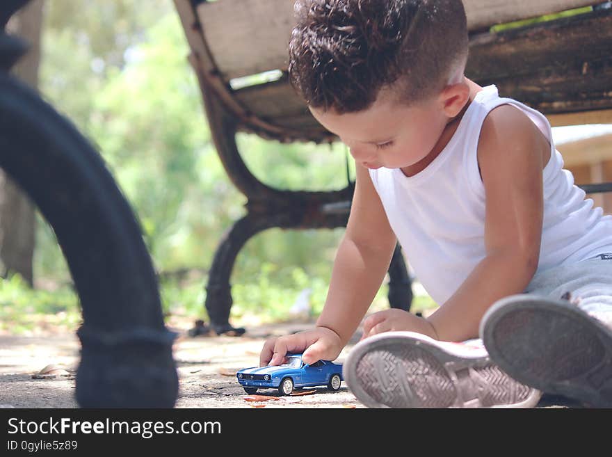 Boy in White Tank Top Playing Blue Coupe Die Cast Near Brown Wooden Bench Chair during Daytime