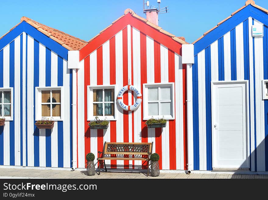 Three red and blue striped beach huts with tiled roofs decorated with a lifebelt and with a bench seat in front, blue sky and sunshine. Three red and blue striped beach huts with tiled roofs decorated with a lifebelt and with a bench seat in front, blue sky and sunshine.