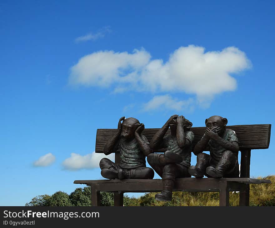 Three little brown monkeys, sculpted from wood, on a wooden bench seat in a park, background blue sky and cloud. The subject could be 'See no evil, hear no evil and speak no evil'. Three little brown monkeys, sculpted from wood, on a wooden bench seat in a park, background blue sky and cloud. The subject could be 'See no evil, hear no evil and speak no evil'.