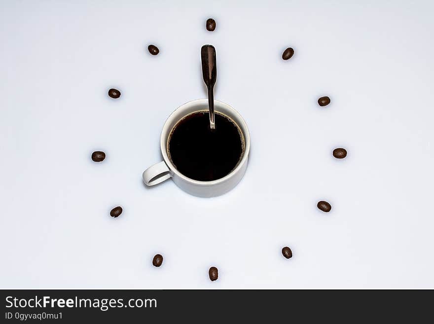 Overhead view of a cup of coffee with a spoon sitting in the cup, surrounding by a circle of evenly spaced coffee beans, on a white background. Overhead view of a cup of coffee with a spoon sitting in the cup, surrounding by a circle of evenly spaced coffee beans, on a white background.