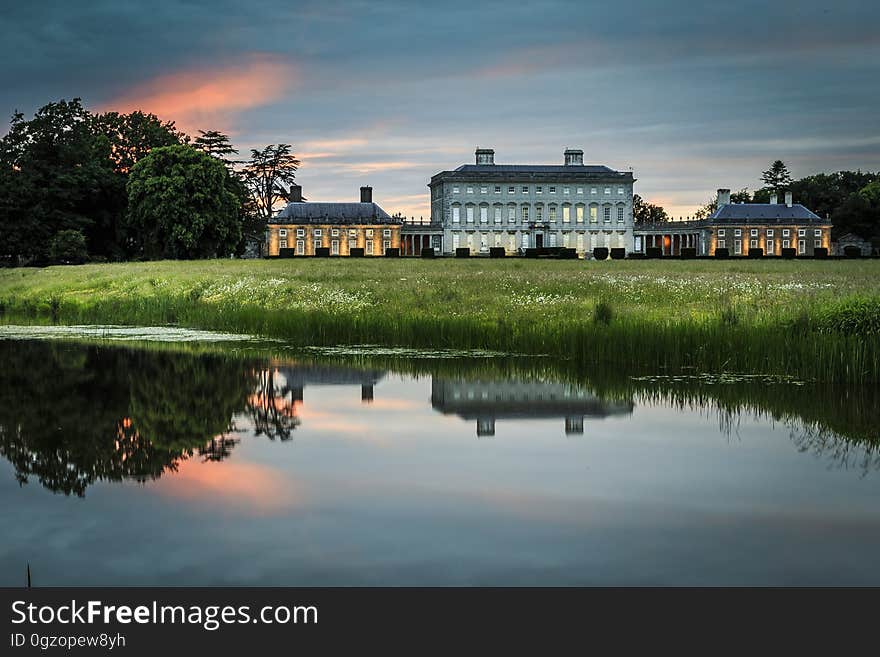 Exterior of estate manor house with green lawn reflecting in waterfront of lake or river at sunset. Exterior of estate manor house with green lawn reflecting in waterfront of lake or river at sunset.