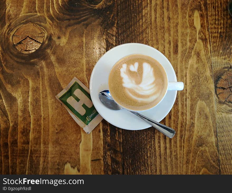 White cup and saucer with coffee or latte with spoon and sugar packet on wooden table. White cup and saucer with coffee or latte with spoon and sugar packet on wooden table.