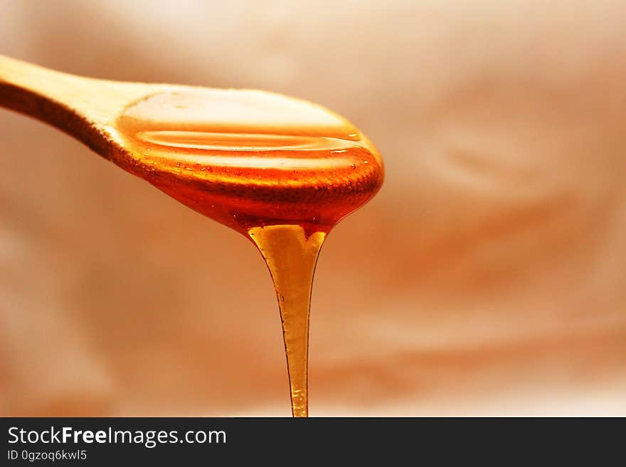 Spoonful of honey with thick sweet liquid flowing over the brim, pale gold background.