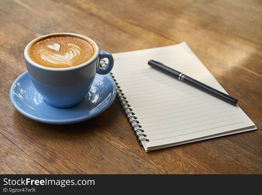 Blue cup and saucer with coffee on wooden table next to blank open notebook and pen. Blue cup and saucer with coffee on wooden table next to blank open notebook and pen.