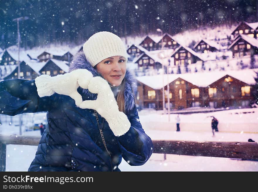 Portrait of woman in winter coat and cap making heart shape with mittens on hands outdoors in snowy town. Portrait of woman in winter coat and cap making heart shape with mittens on hands outdoors in snowy town.