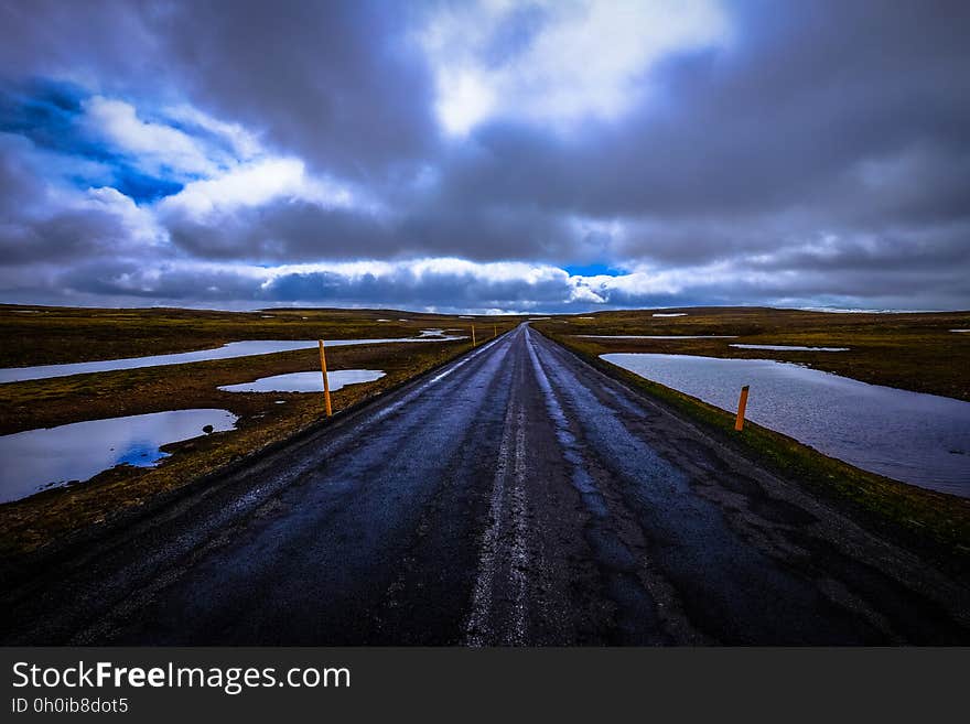 Long straight road with center markings through countryside with fields and a river and pools of flood water, rain clouds overhead. Long straight road with center markings through countryside with fields and a river and pools of flood water, rain clouds overhead.