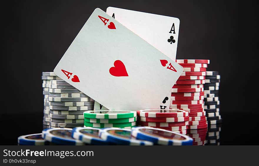 Ace of hearts and ace of clubs leaning against large piles of red, gray, green and blue chips, dark background. Ace of hearts and ace of clubs leaning against large piles of red, gray, green and blue chips, dark background.