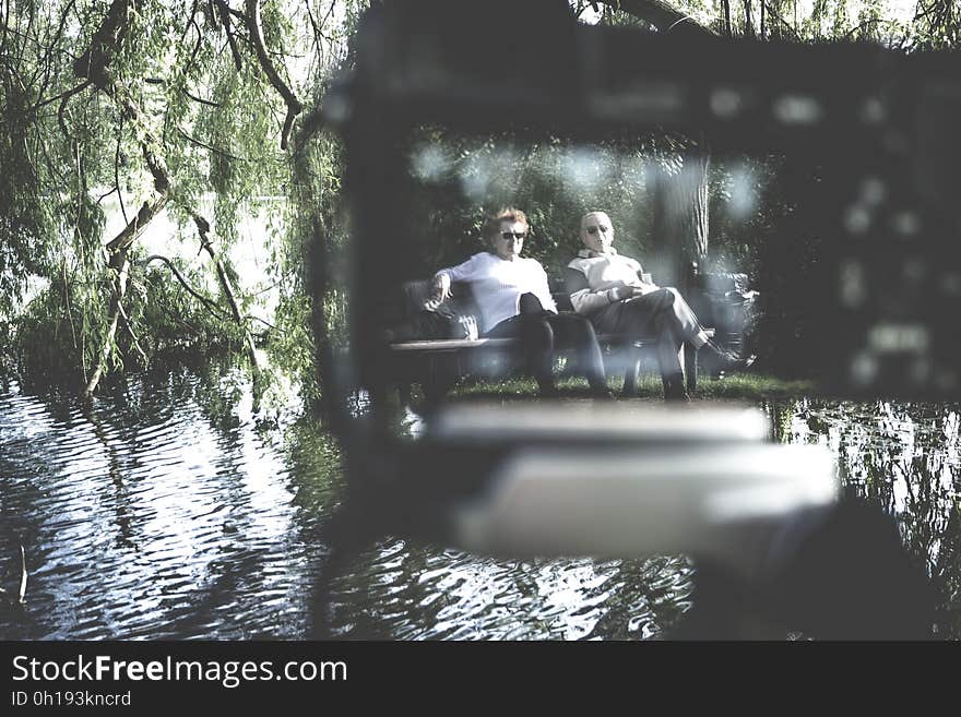 Men sitting on park bench next to pond on sunny day. Men sitting on park bench next to pond on sunny day.