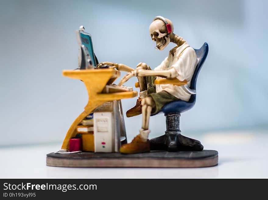 Skeleton (sculpture) wearing headphones sitting in a black swivel chair working the keyboard of a computer on a yellow desk, white and pale blue background. Concept of computers killing us. Skeleton (sculpture) wearing headphones sitting in a black swivel chair working the keyboard of a computer on a yellow desk, white and pale blue background. Concept of computers killing us.